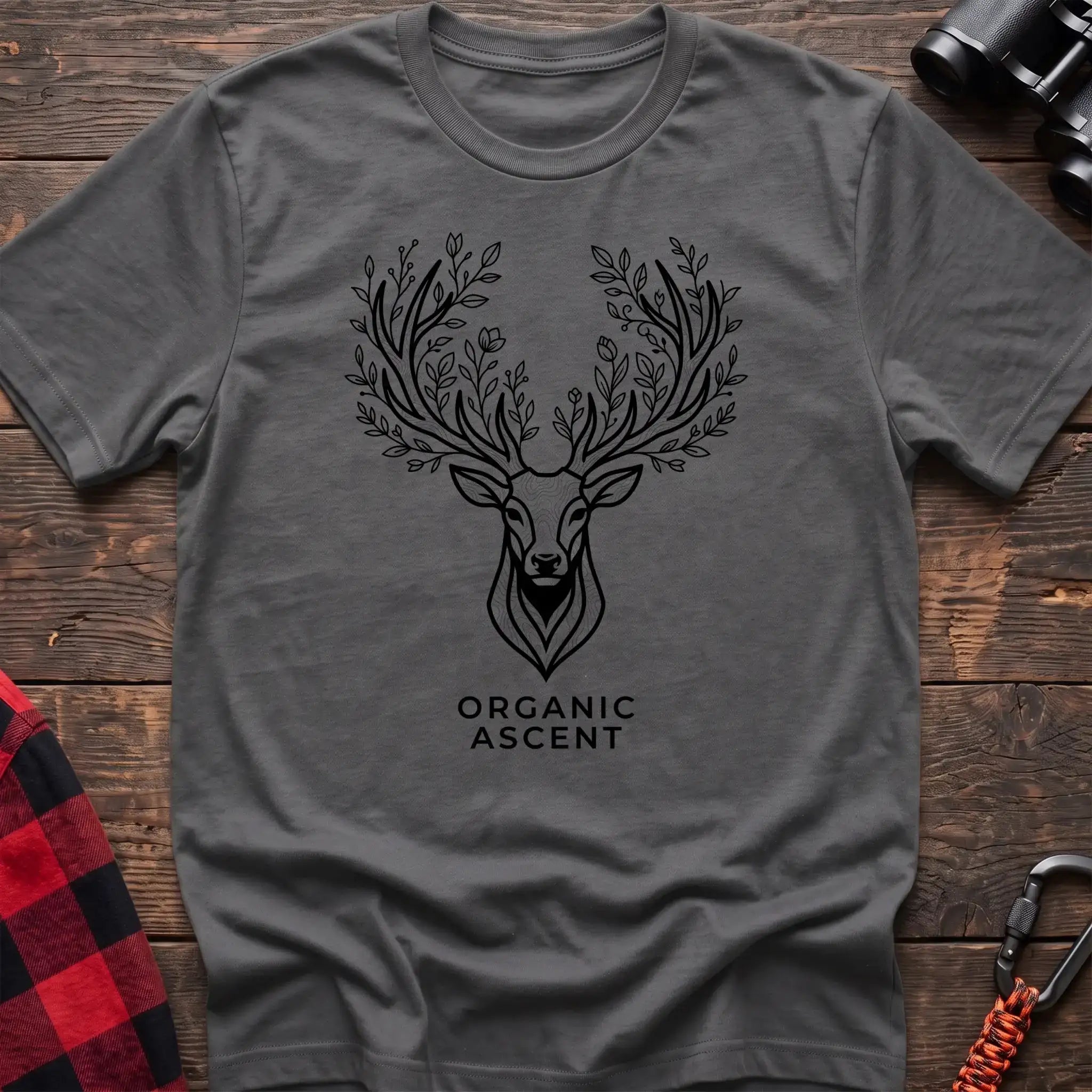 Graphite heather t-shirt with a black line drawing of a deer’s head whose antlers are formed from leafy branches and flowers, with the text “ORGANIC ASCENT” printed below, laid flat on a wooden surface.