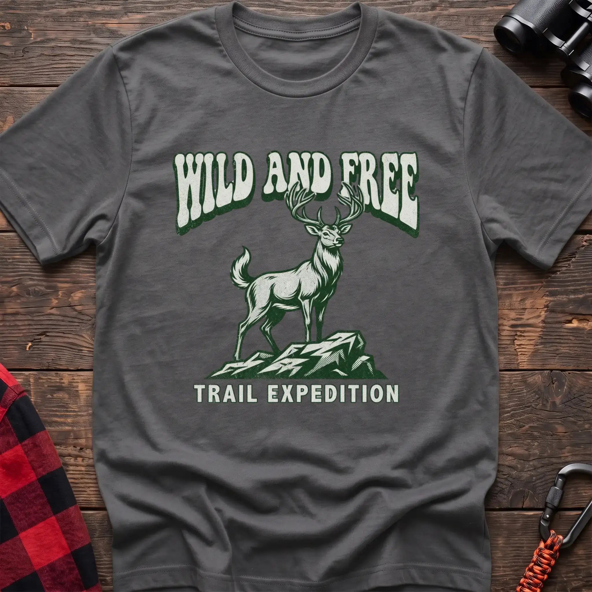 Graphite heather t-shirt with a rustic graphic of a deer standing on a rocky outcrop, with the arched text “WILD AND FREE” above and “TRAIL EXPEDITION” below, laid flat on a wooden surface.