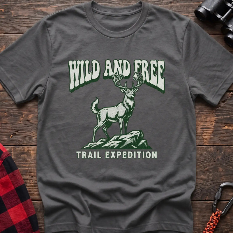 Graphite heather t-shirt with a rustic graphic of a deer standing on a rocky outcrop, with the arched text “WILD AND FREE” above and “TRAIL EXPEDITION” below, laid flat on a wooden surface.