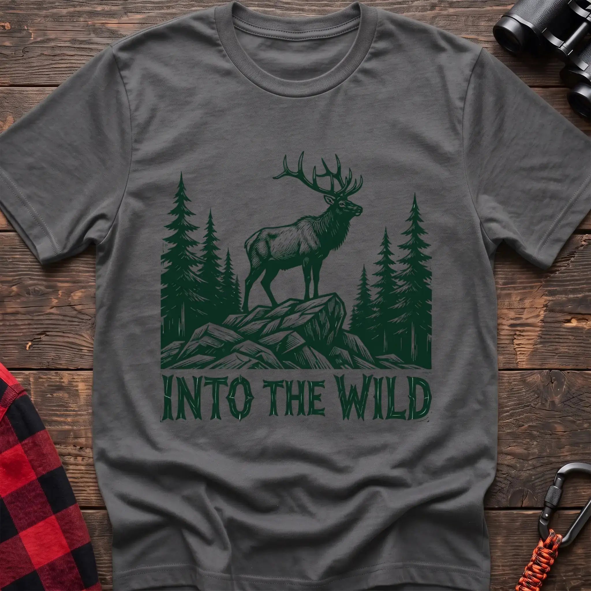 Graphite heather t-shirt with a dark green illustration of an elk standing on a rocky outcrop among pine trees, with the text “INTO THE WILD” printed below, laid flat on a wooden surface.