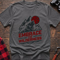 Graphite heather t-shirt with a dark graphic of a howling wolf against mountain peaks and a red sun, with the bold red text “EMBRACE THE WILDERNESS” below, laid flat on a wooden surface.