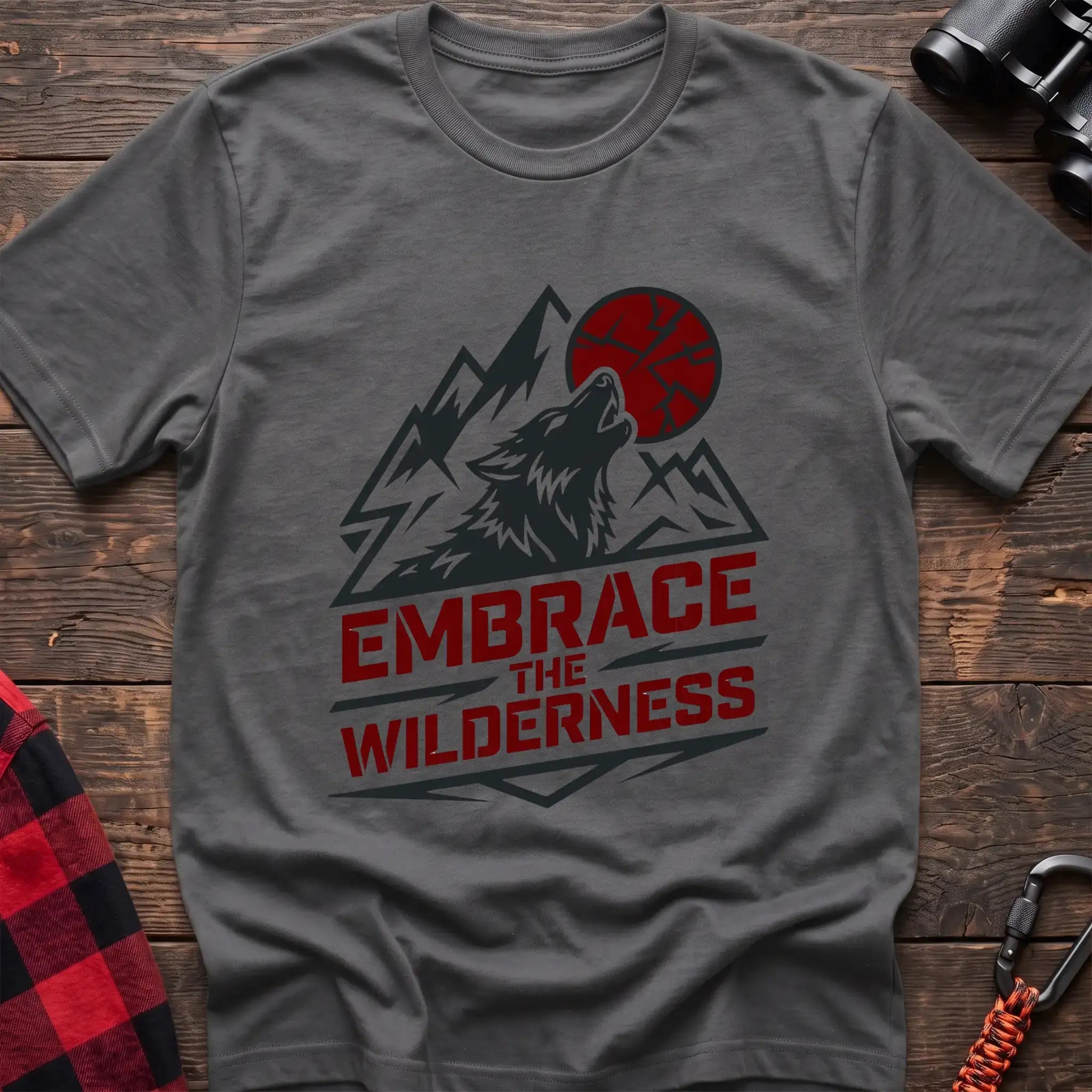 Graphite heather t-shirt with a dark graphic of a howling wolf against mountain peaks and a red sun, with the bold red text “EMBRACE THE WILDERNESS” below, laid flat on a wooden surface.