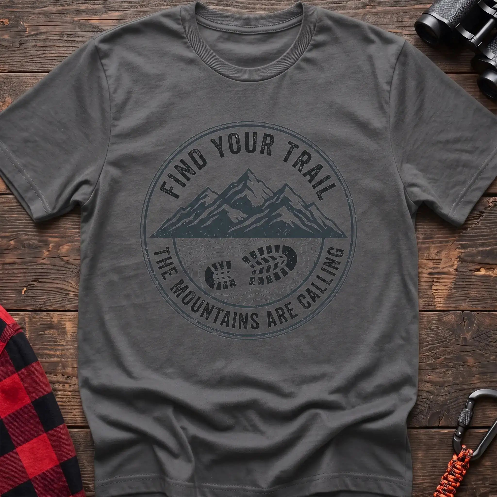 Graphite heather t-shirt with a circular dark gray badge design featuring mountain peaks above a hiking boot print on topographic lines, with the text “FIND YOUR TRAIL” and “THE MOUNTAINS ARE CALLING,” laid flat on a wooden surface.