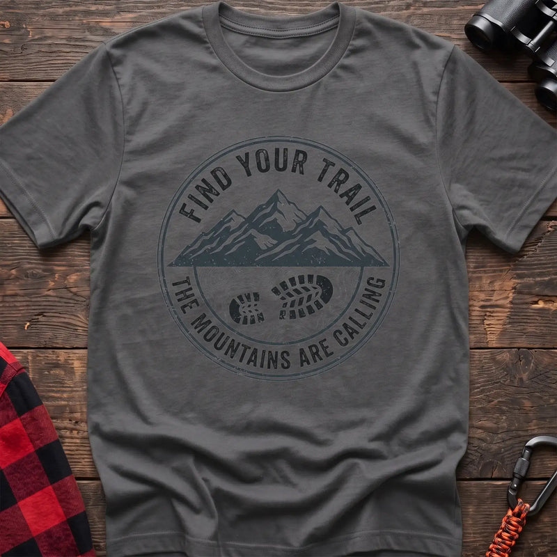 Graphite heather t-shirt with a circular dark gray badge design featuring mountain peaks above a hiking boot print on topographic lines, with the text “FIND YOUR TRAIL” and “THE MOUNTAINS ARE CALLING,” laid flat on a wooden surface.