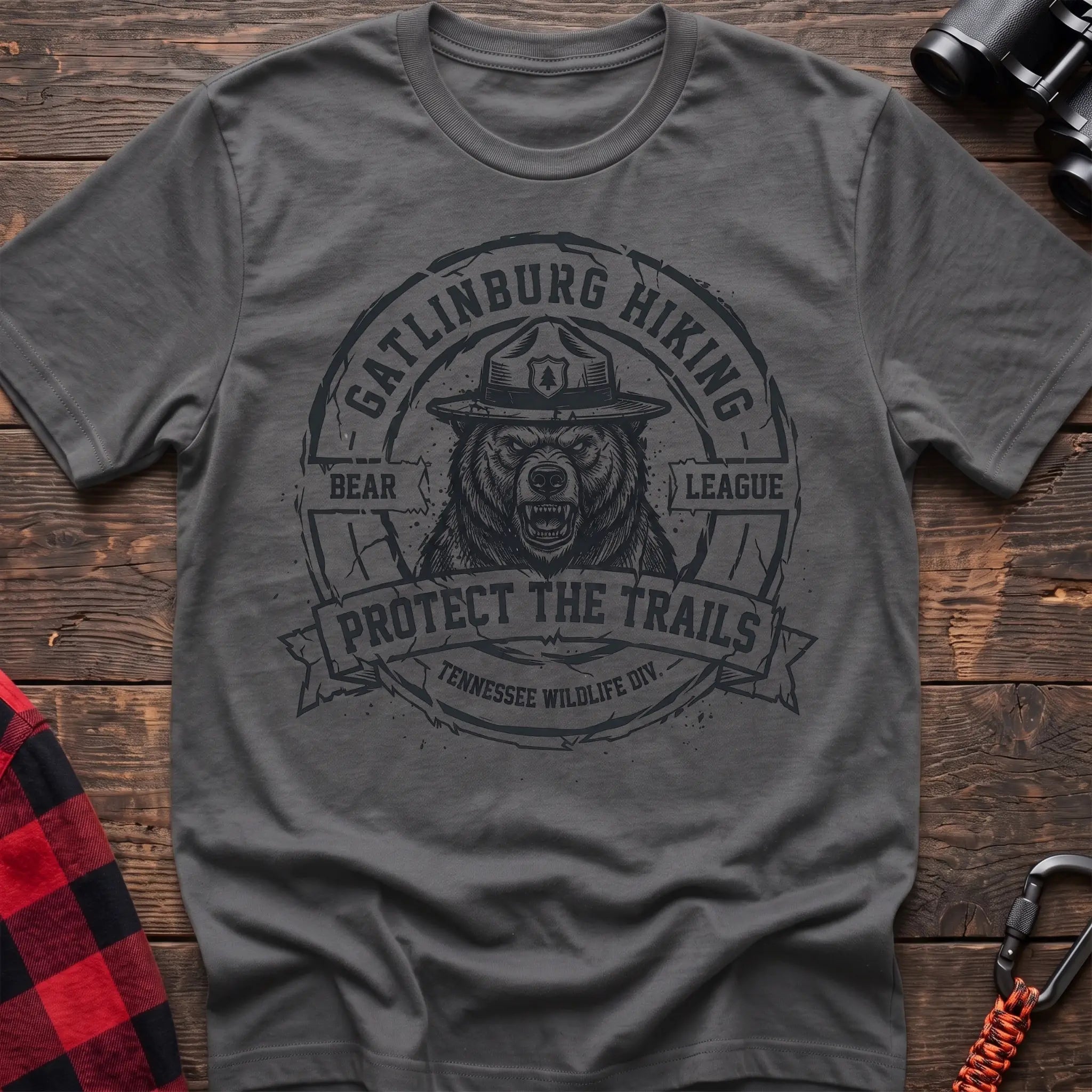 Graphite heather cotton t-shirt with vintage Gatlinburg Hiking Bear League graphic of a roaring bear in a park ranger hat, Protect the Trails Tennessee Wildlife Division badge, flat lay on rustic wood with hiking gear