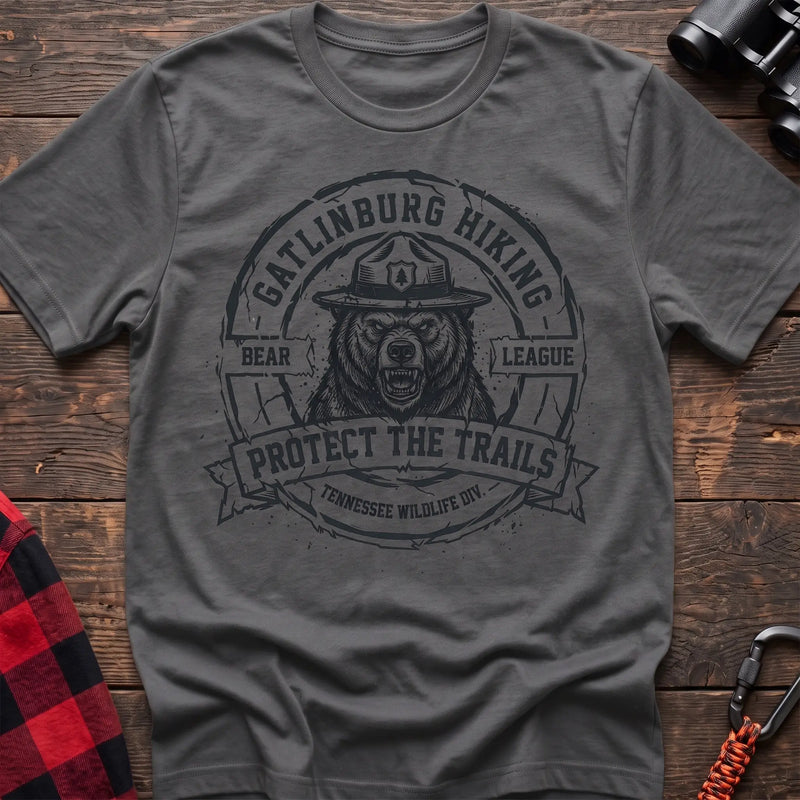 Graphite heather cotton t-shirt with vintage Gatlinburg Hiking Bear League graphic of a roaring bear in a park ranger hat, Protect the Trails Tennessee Wildlife Division badge, flat lay on rustic wood with hiking gear