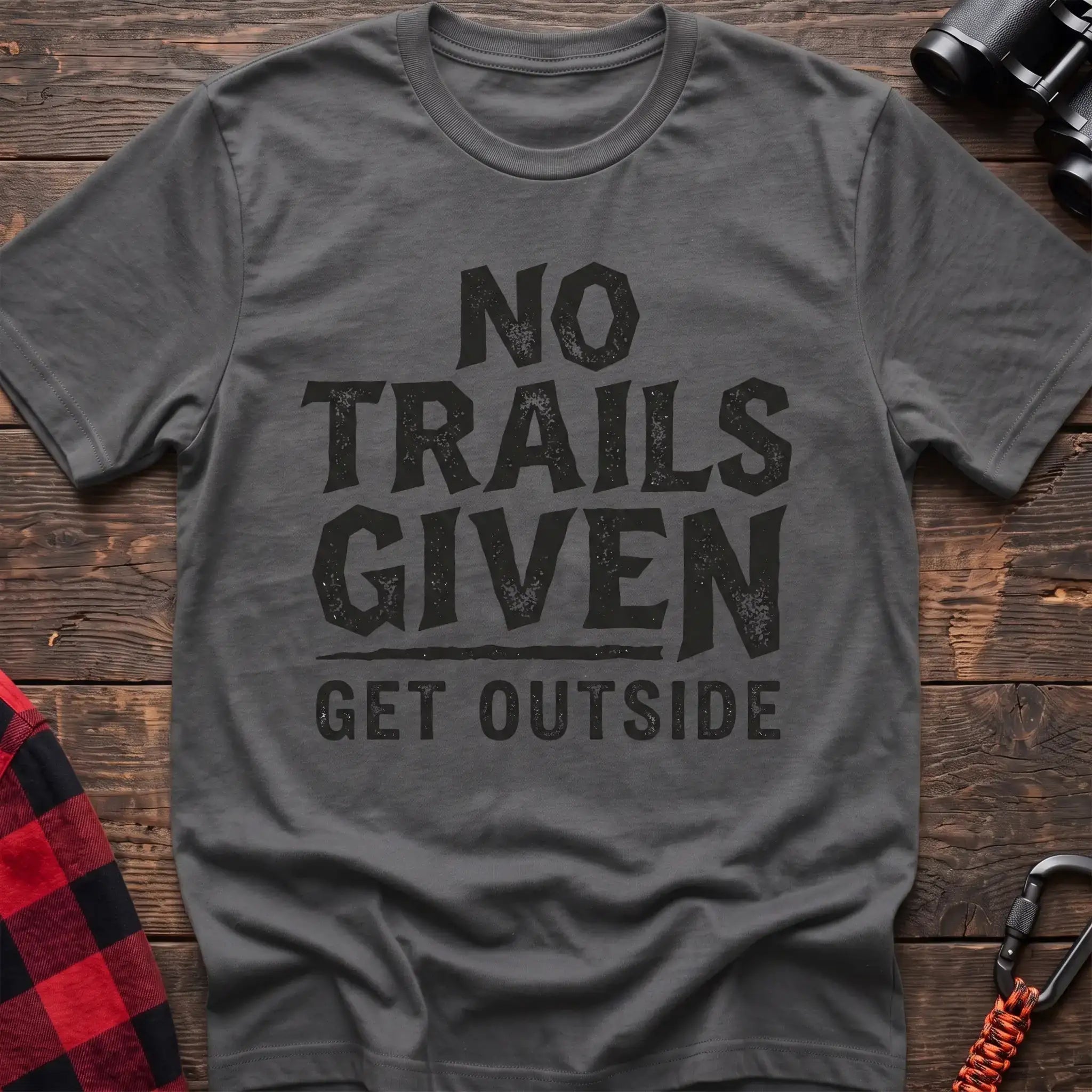 Graphite heather t-shirt with distressed black text reading “NO TRAILS GIVEN” and “GET OUTSIDE” below it, laid flat on a wooden surface.