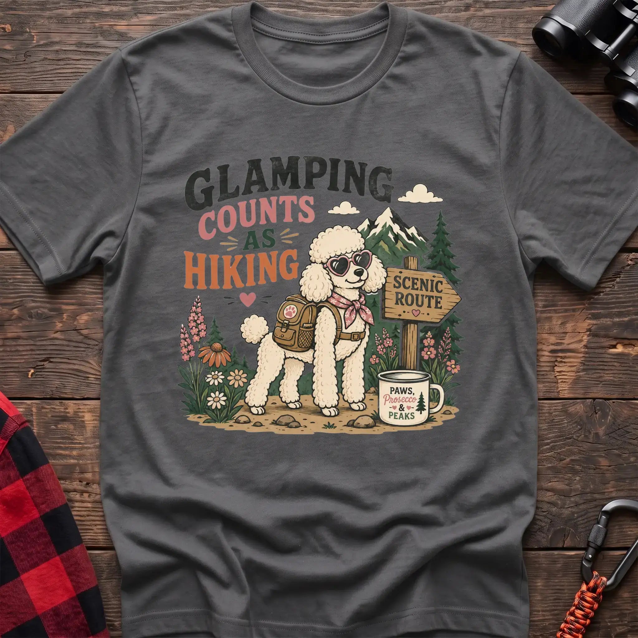 Graphite heather t-shirt with a colorful illustration of a poodle in sunglasses and a backpack on a scenic trail, with the text “GLAMPING COUNTS AS HIKING,” laid flat on a wooden surface.