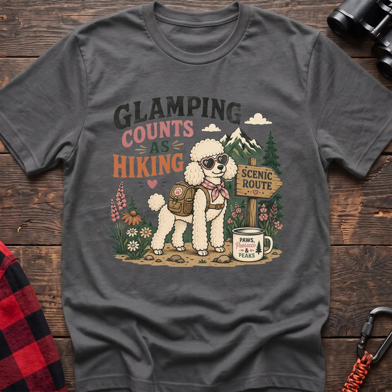 Graphite heather t-shirt with a colorful illustration of a poodle in sunglasses and a backpack on a scenic trail, with the text “GLAMPING COUNTS AS HIKING,” laid flat on a wooden surface.