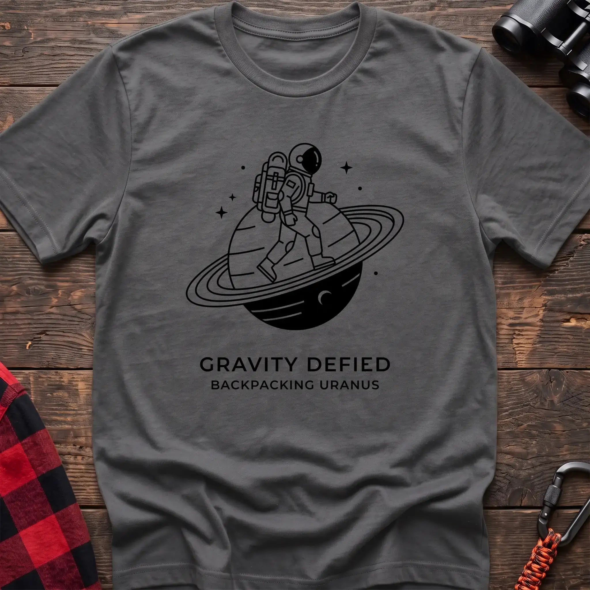 Graphite heather crew-neck t-shirt with black line-art graphic of an astronaut backpacking across a ringed planet surrounded by stars, with the text “GRAVITY DEFIED / BACKPACKING URANUS” printed below, laid flat on a wooden surface.