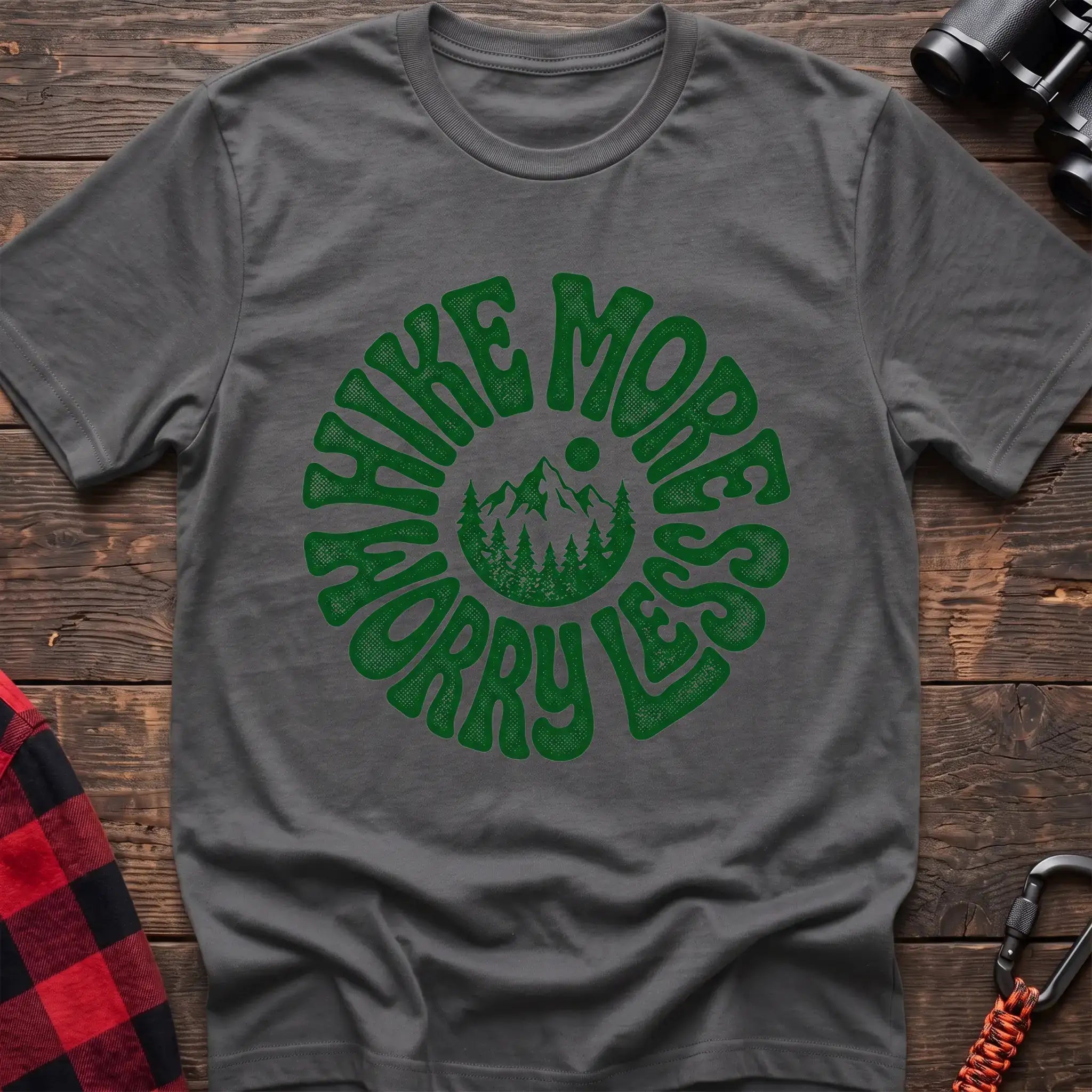 Graphite heather t-shirt with a circular green design featuring mountains and pine trees in the center, surrounded by the text “HIKE MORE WORRY LESS,” laid flat on a wooden surface.