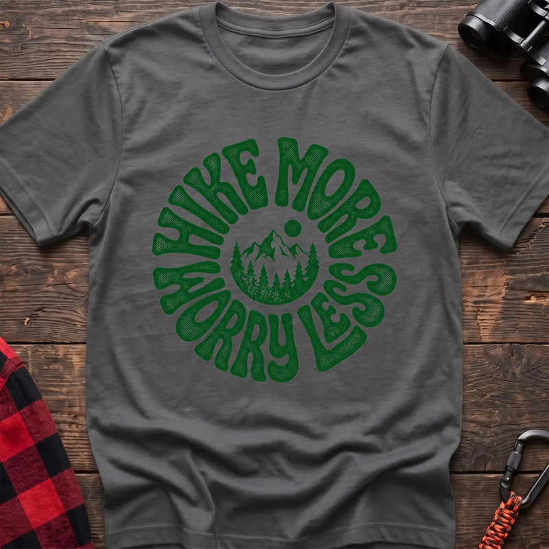 Graphite heather t-shirt with a circular green design featuring mountains and pine trees in the center, surrounded by the text “HIKE MORE WORRY LESS,” laid flat on a wooden surface.