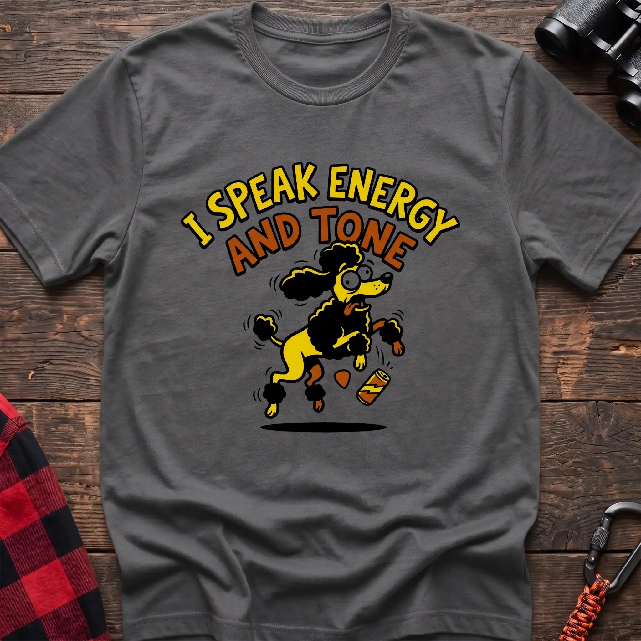 Graphite heather t-shirt with a cartoon yellow and black poodle jumping excitedly next to a falling energy drink can, with the arched text “I SPEAK ENERGY AND TONE,” laid flat on a wooden surface.