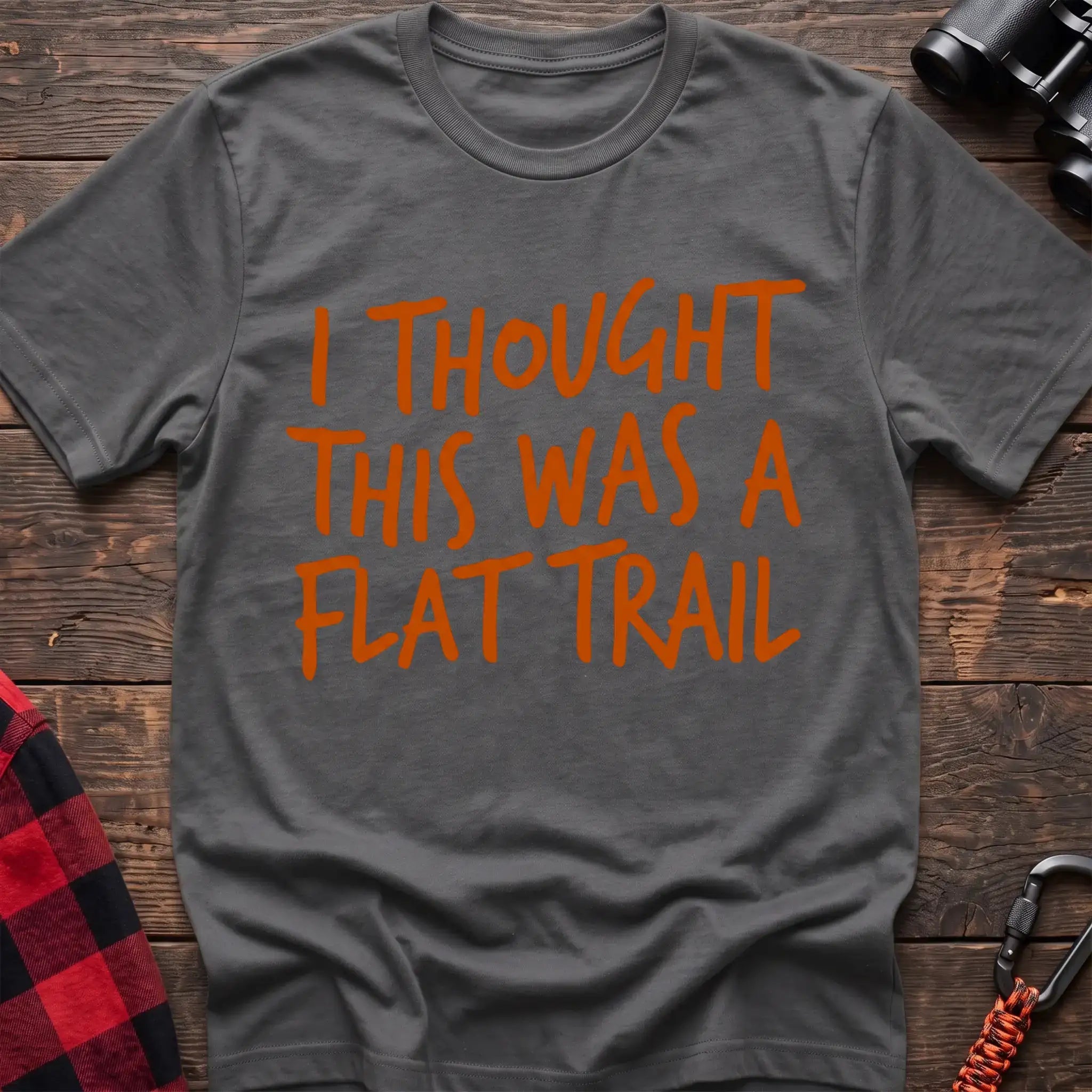 Graphite heather t-shirt with bold orange handwritten-style text reading “I THOUGHT THIS WAS A FLAT TRAIL,” laid flat on a wooden surface.