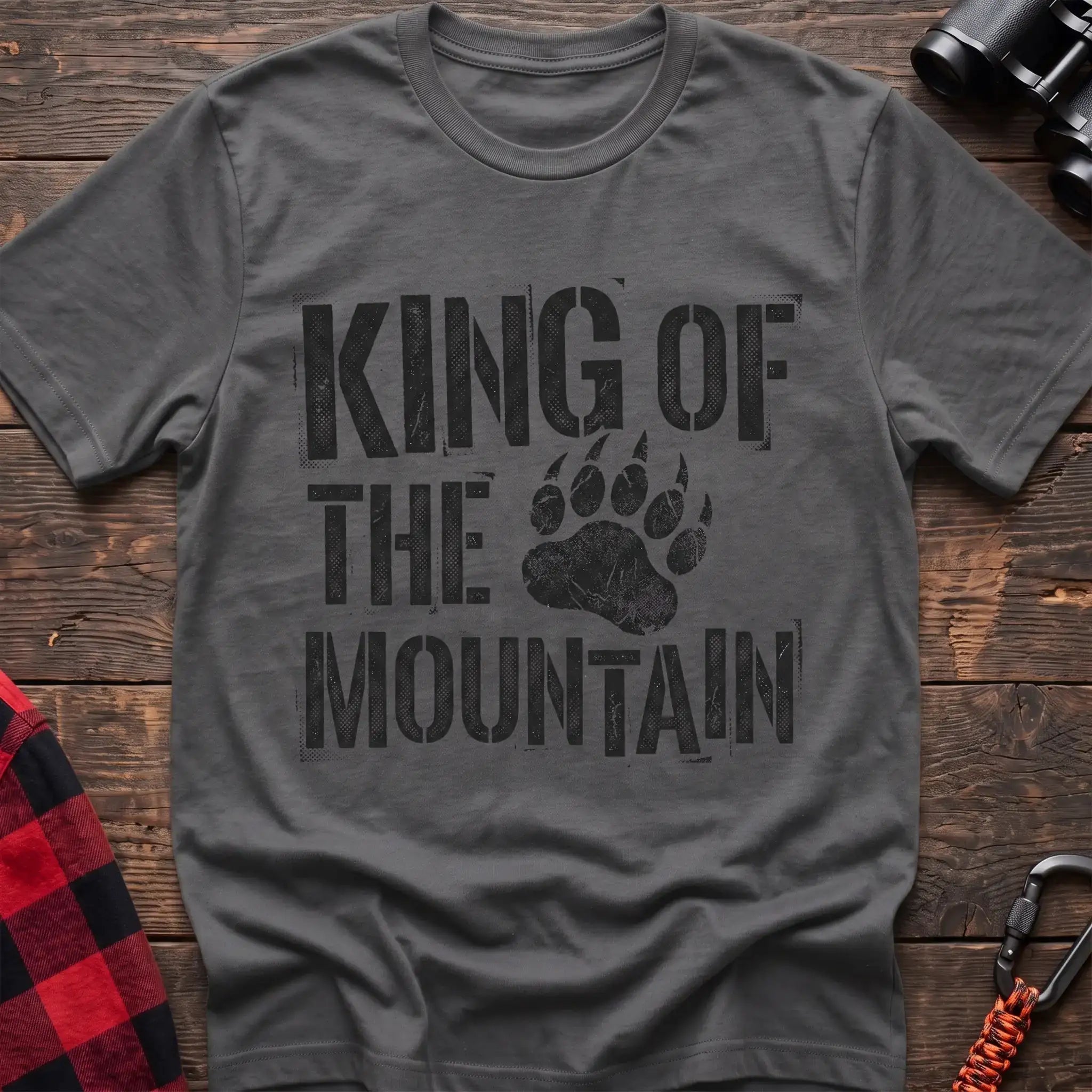 Graphite heather t-shirt with distressed black text reading “KING OF THE MOUNTAIN” and a large bear paw print integrated into the design, laid flat on a wooden surface.