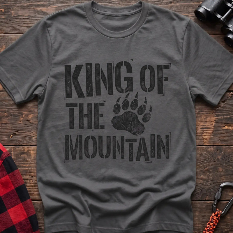 Graphite heather t-shirt with distressed black text reading “KING OF THE MOUNTAIN” and a large bear paw print integrated into the design, laid flat on a wooden surface.