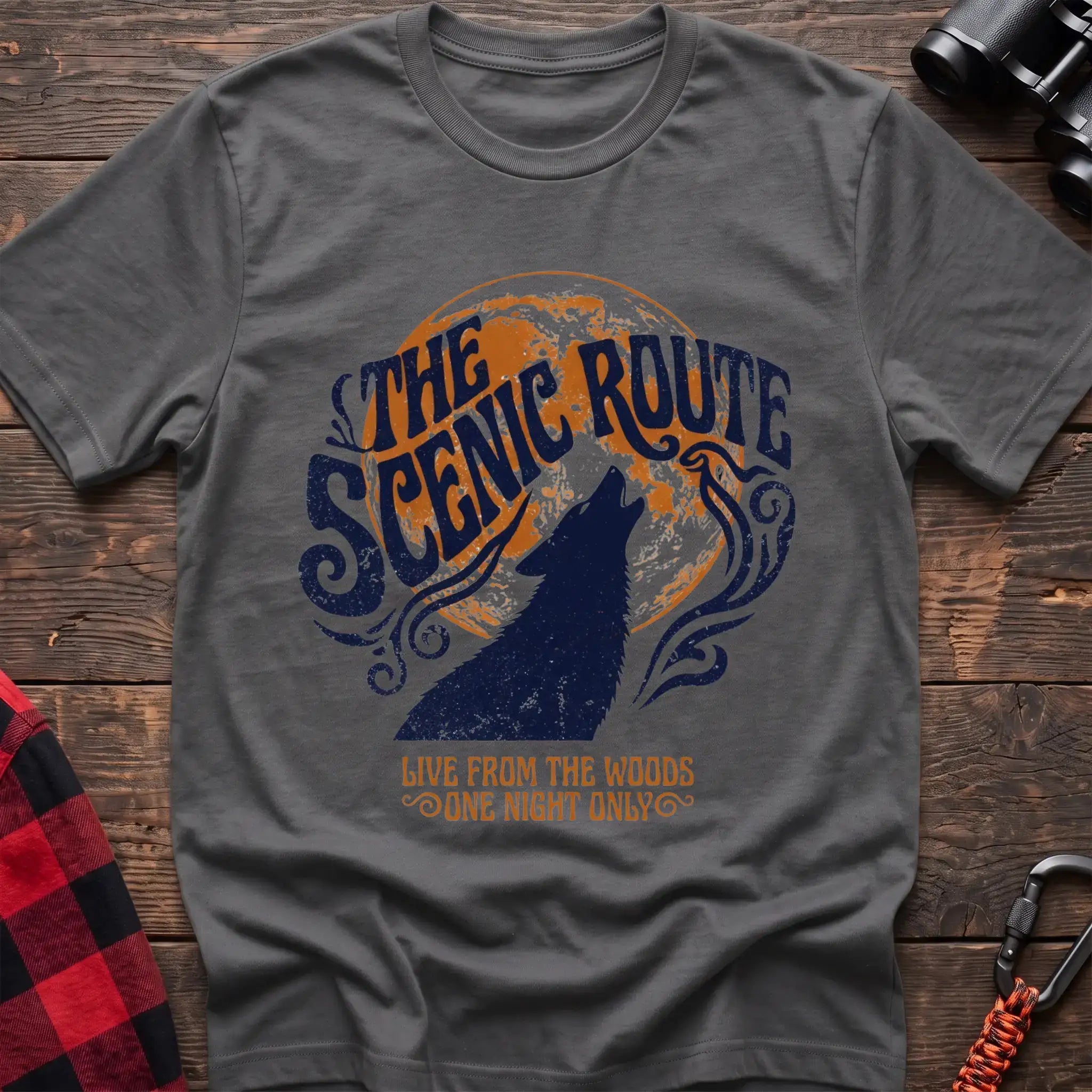 Graphite heather t-shirt with a dark blue howling wolf silhouette against an orange circle, with the arched text “THE SCENIC ROUTE” above and “LIVE FROM THE WOODS / ONE NIGHT ONLY” below, laid flat on a wooden surface.