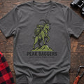 Graphite heather t-shirt with an illustration of a mountain goat wearing a backpack standing on a peak, with the text “PEAK BAGGERS” and “LIGHT LOADS, STEEP CLIMBS,” laid flat on a wooden surface.