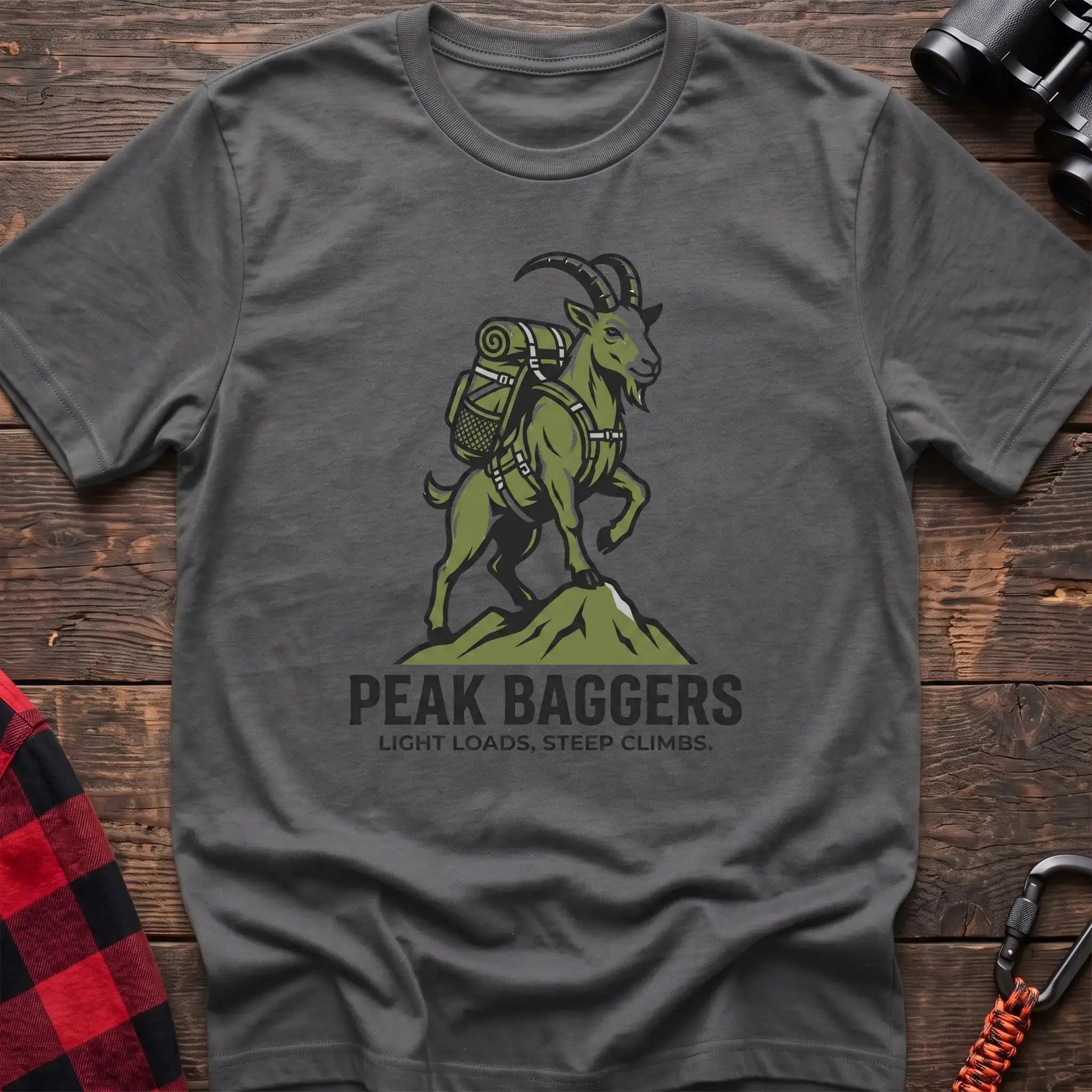 Graphite heather t-shirt with an illustration of a mountain goat wearing a backpack standing on a peak, with the text “PEAK BAGGERS” and “LIGHT LOADS, STEEP CLIMBS,” laid flat on a wooden surface.
