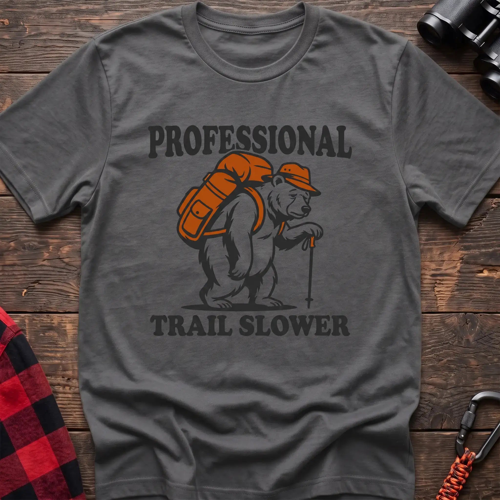 Graphite heather t-shirt with a cartoon polar bear wearing a backpack and hat while holding a hiking stick, with the text “PROFESSIONAL TRAIL SLOWER,” laid flat on a wooden surface.