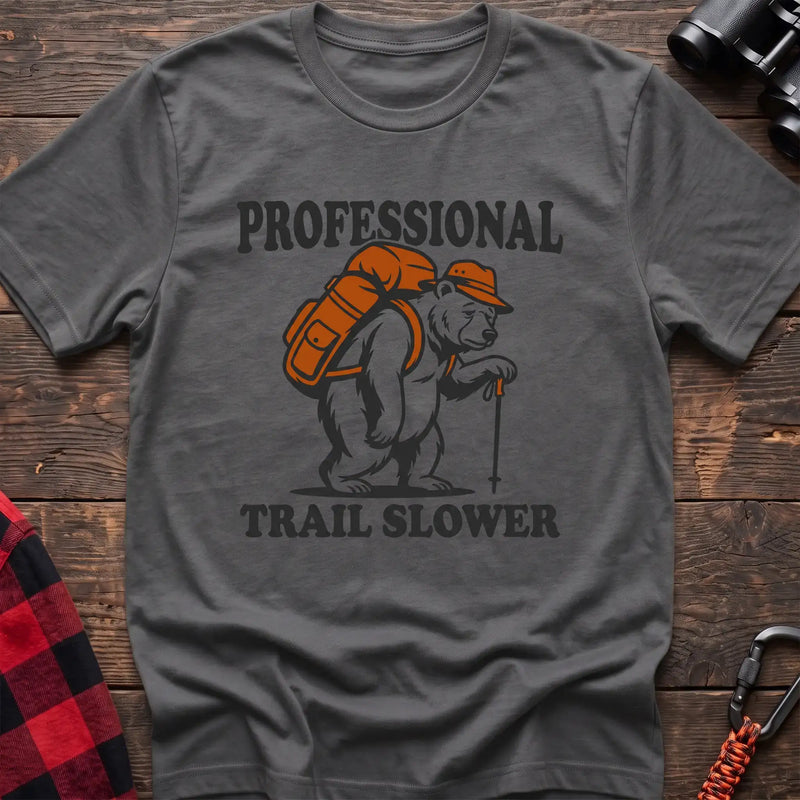 Graphite heather t-shirt with a cartoon polar bear wearing a backpack and hat while holding a hiking stick, with the text “PROFESSIONAL TRAIL SLOWER,” laid flat on a wooden surface.