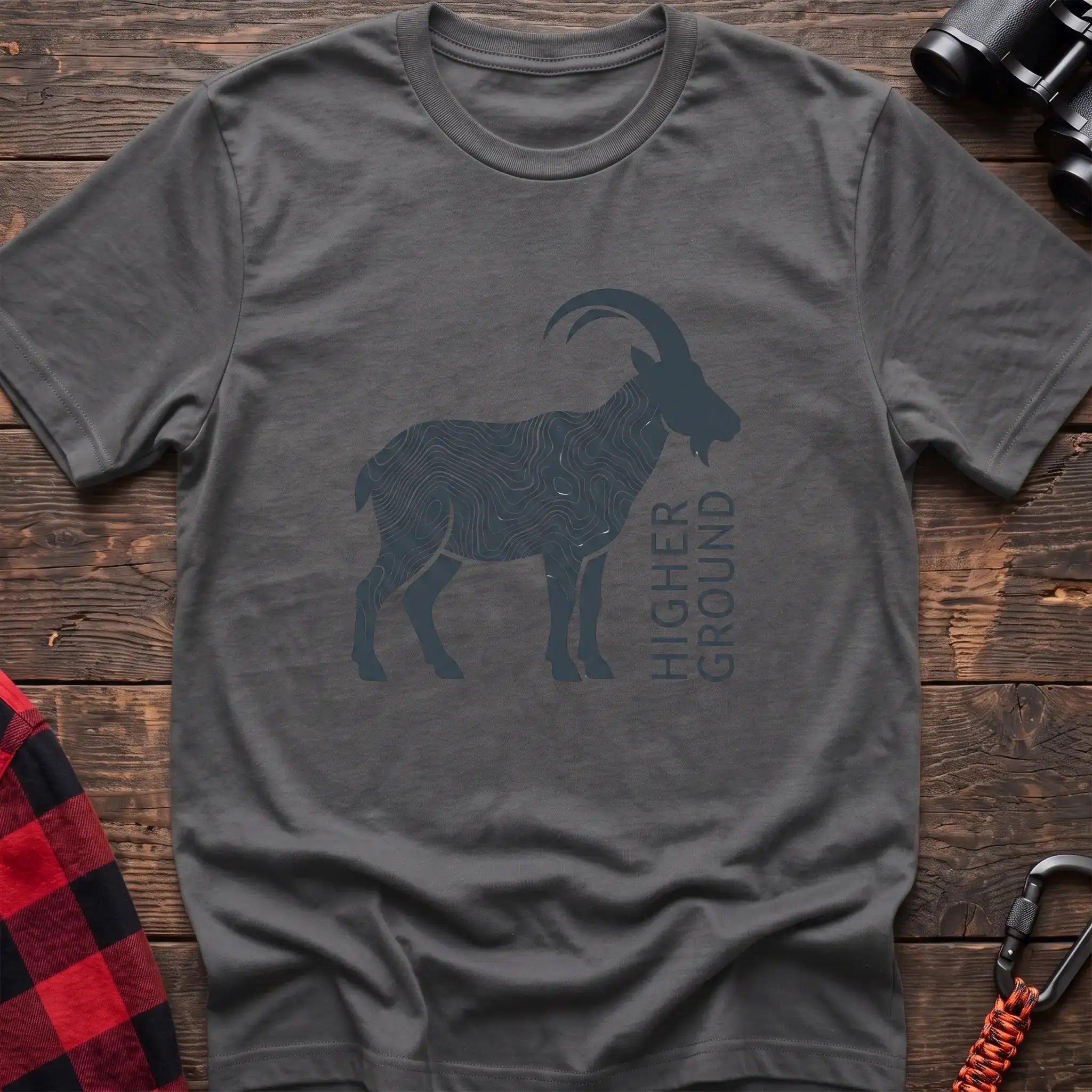 Graphite heather t-shirt with a dark silhouette of a mountain goat filled with topographic contour lines, and the vertical text “HIGHER GROUND” printed beside it, laid flat on a wooden surface.