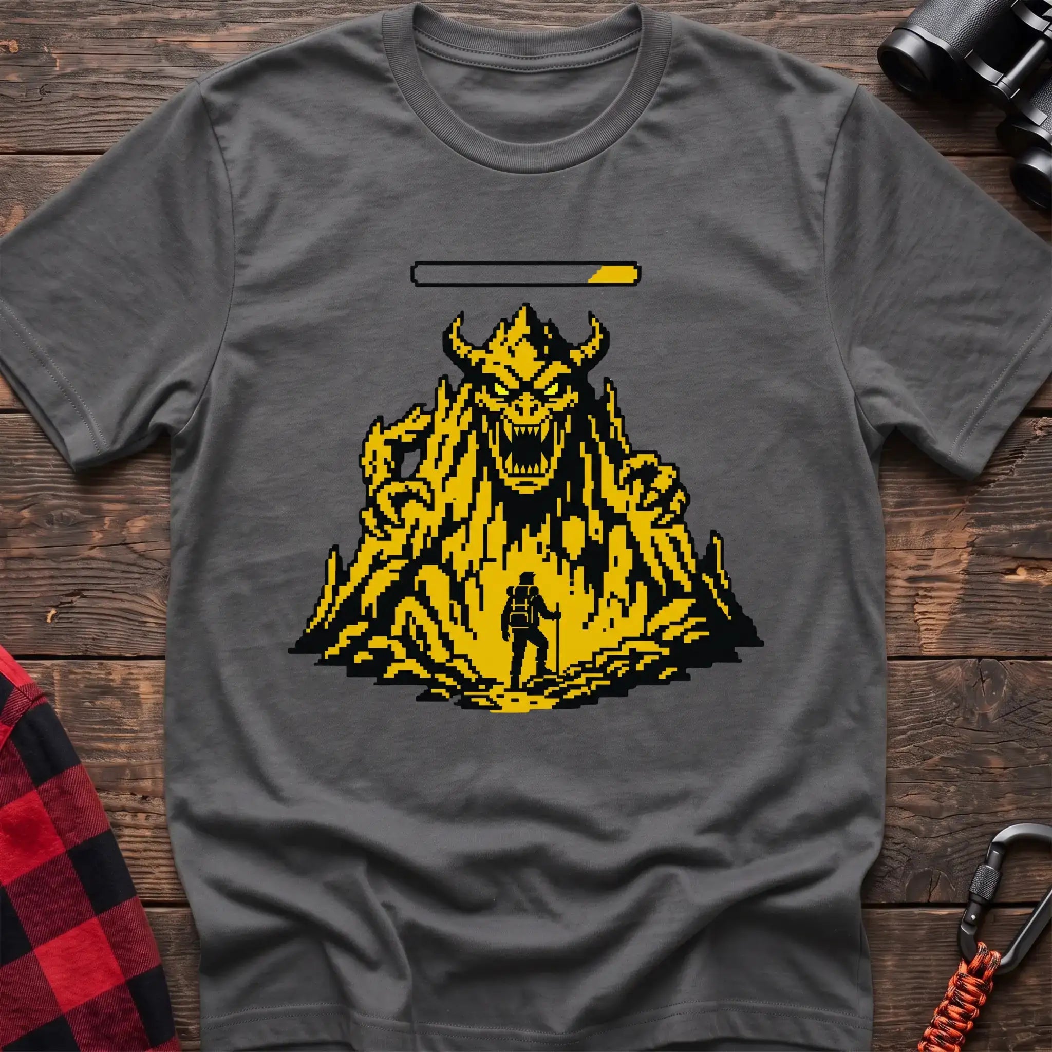 Graphite heather t-shirt with a pixel-art illustration of a hiker facing a large horned mountain monster with a health bar above it, laid flat on a wooden surface.