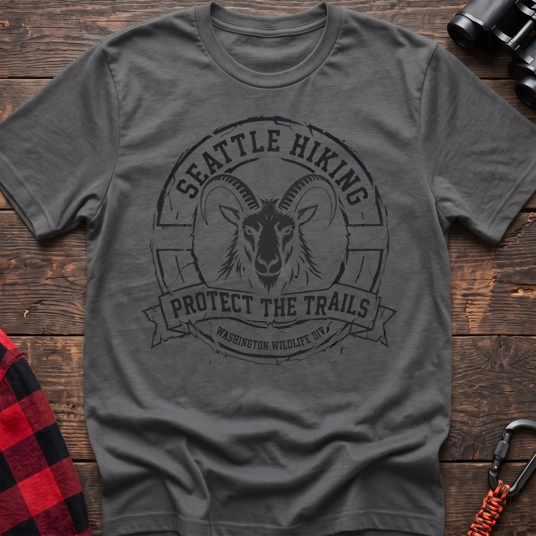 Graphite heather cotton t-shirt with vintage Seattle Hiking graphic of a mountain goat in a circular badge, Protect the Trails Washington Wildlife Division design, flat lay on rustic wood with hiking gear