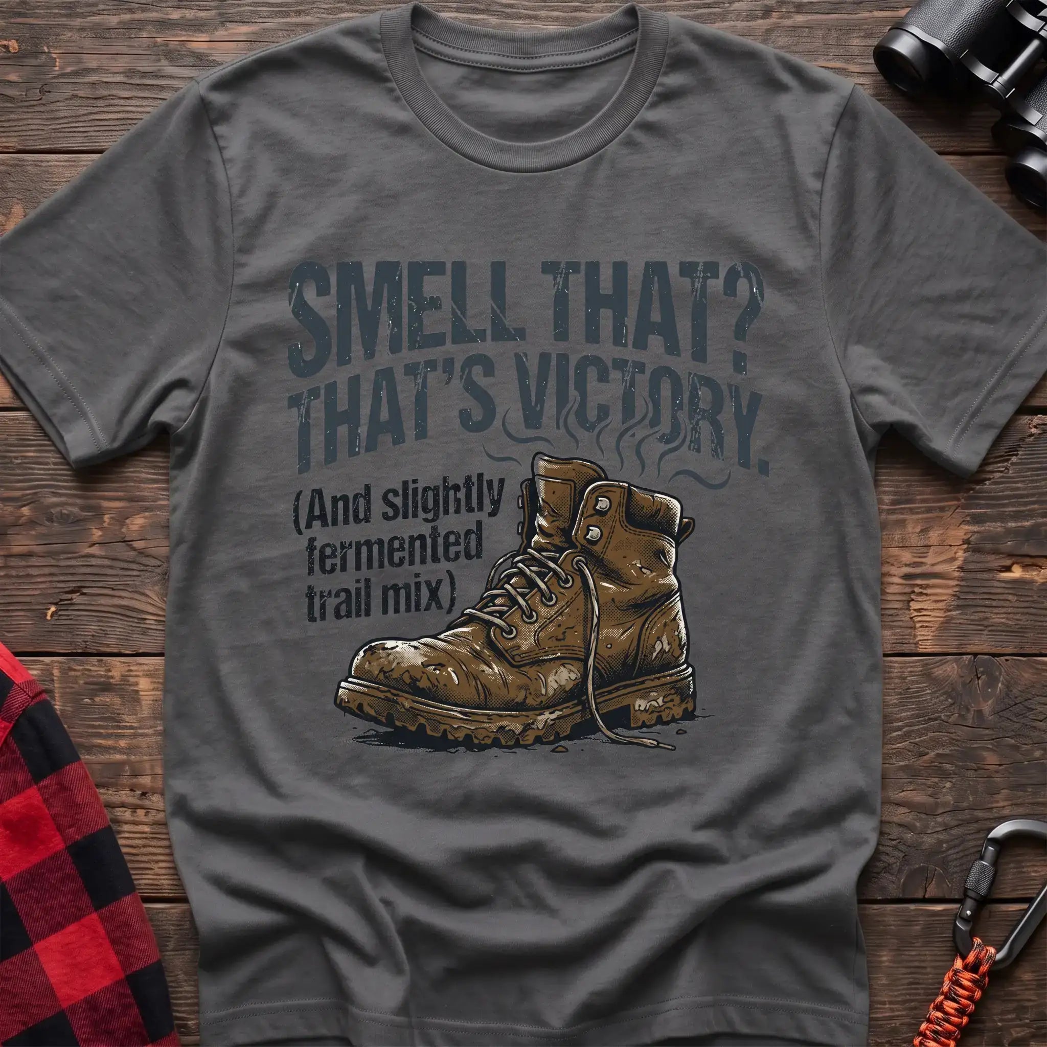 Graphite heather t-shirt with an illustration of a dirty hiking boot with rising steam lines, and the text “SMELL THAT? THAT’S VICTORY. (And slightly fermented trail mix),” laid flat on a wooden surface