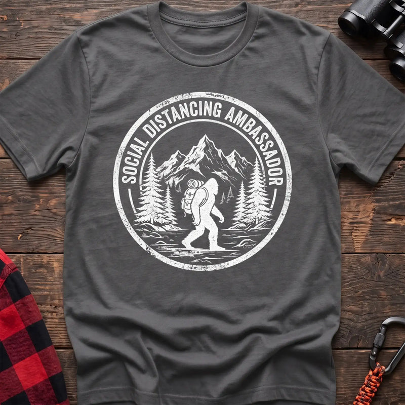 Graphite heather t-shirt with white circular badge graphic of Bigfoot hiking with a backpack through mountains and the text Social Distancing Ambassador