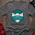 Graphite heather t-shirt with a hexagonal illustration of a person meditating by a mountain lake, with the text “SOLITUDE SEEKER’S GUILD” and “Finding Peace One Hidden Trail at a Time,” laid flat on a wooden surface.