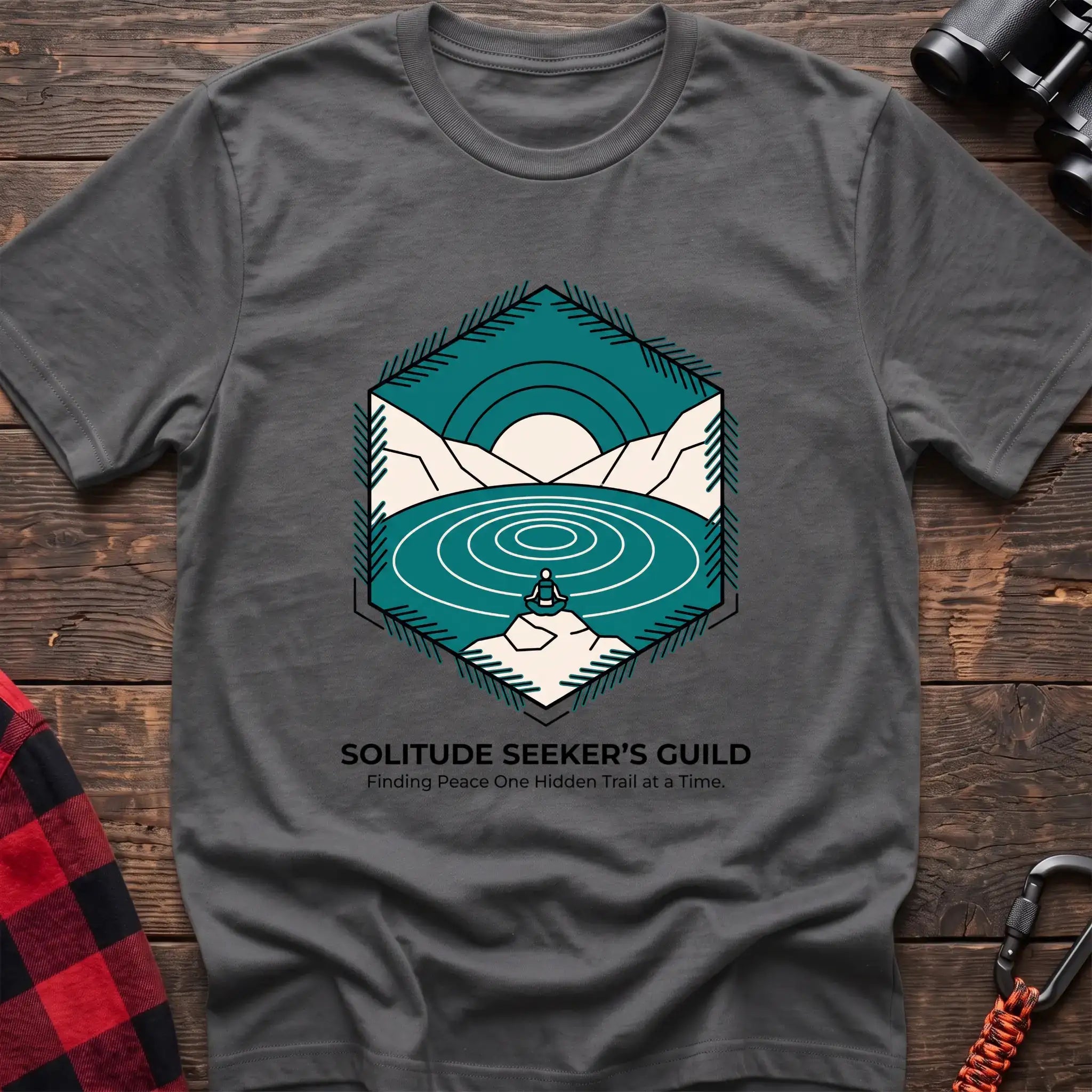 Graphite heather t-shirt with a hexagonal illustration of a person meditating by a mountain lake, with the text “SOLITUDE SEEKER’S GUILD” and “Finding Peace One Hidden Trail at a Time,” laid flat on a wooden surface.