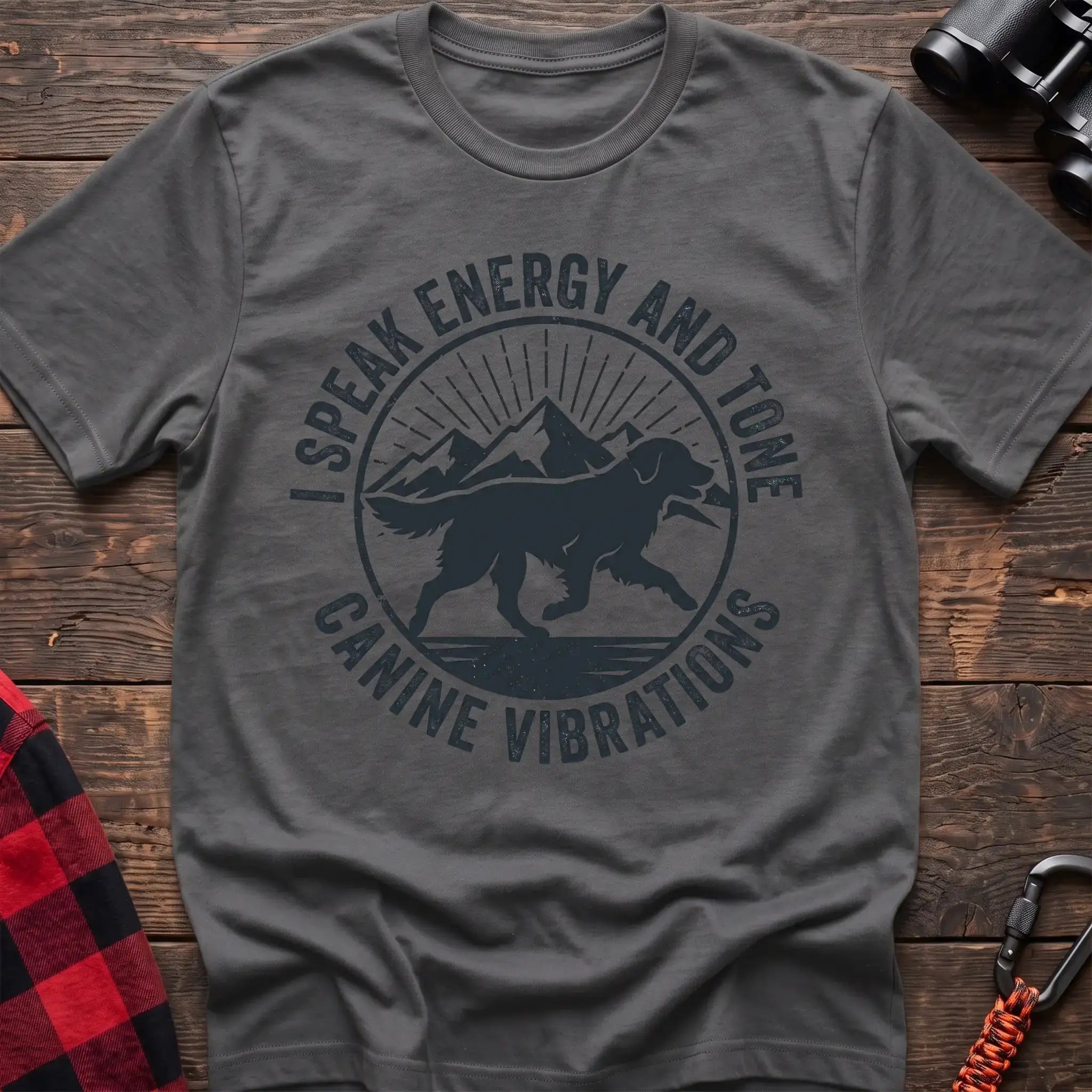 Graphite heather t-shirt with a circular dark gray badge design featuring a running dog carrying a stick against mountain peaks and sun rays, with the text “I SPEAK ENERGY AND TONE” and “CANINE VIBRATIONS,” laid flat on a wooden surface.