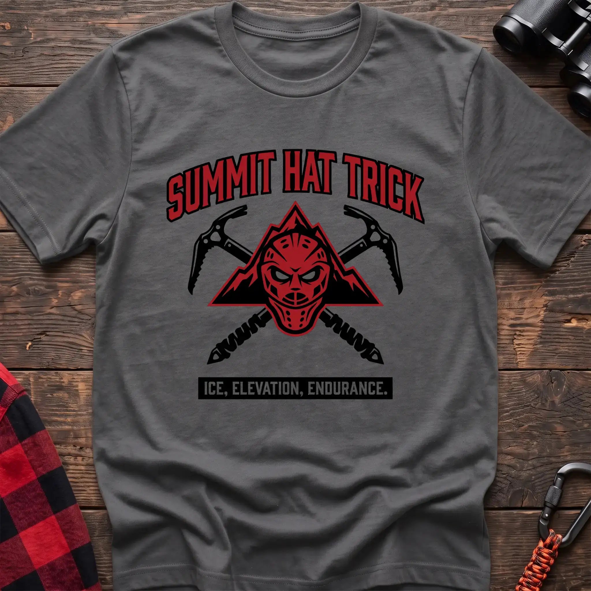 Graphite heather t-shirt with a red mountain-themed goalie mask and crossed ice axes, with the text “SUMMIT HAT TRICK” and “ICE, ELEVATION, ENDURANCE,” laid flat on a wooden surface.