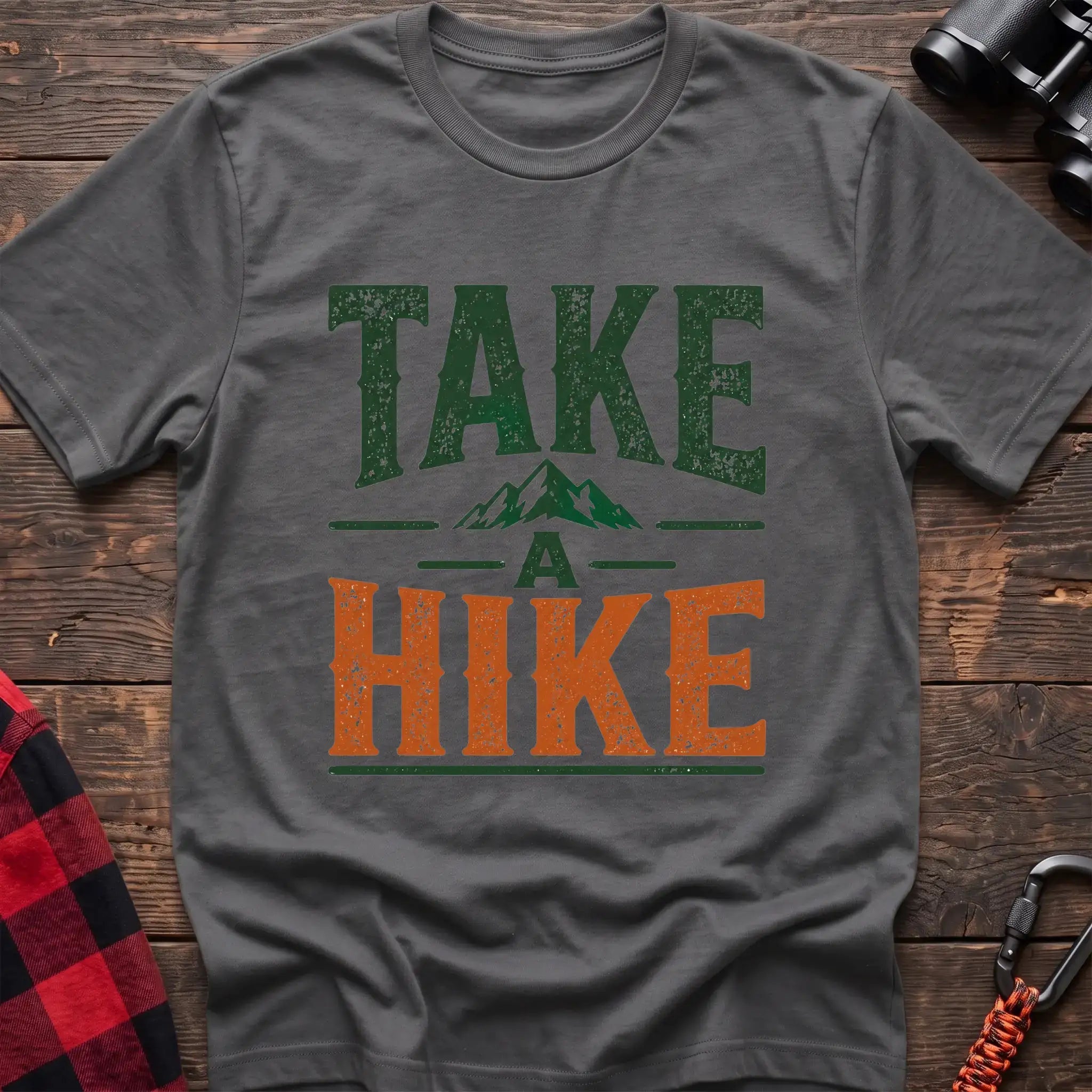 Graphite heather t-shirt with distressed green and orange text reading “TAKE A HIKE” with a small mountain graphic between the words, laid flat on a wooden surface.