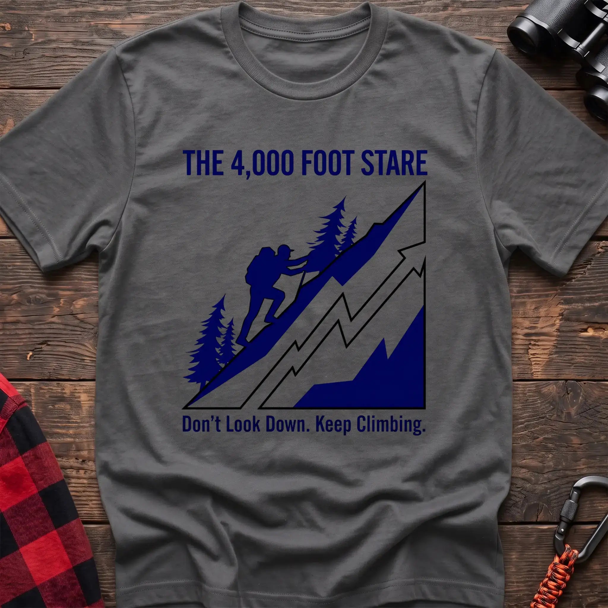 Graphite heather t-shirt with a blue illustration of a hiker climbing a steep mountain, with the text “THE 4,000 FOOT STARE” and “Don’t Look Down. Keep Climbing,” laid flat on a wooden surface.