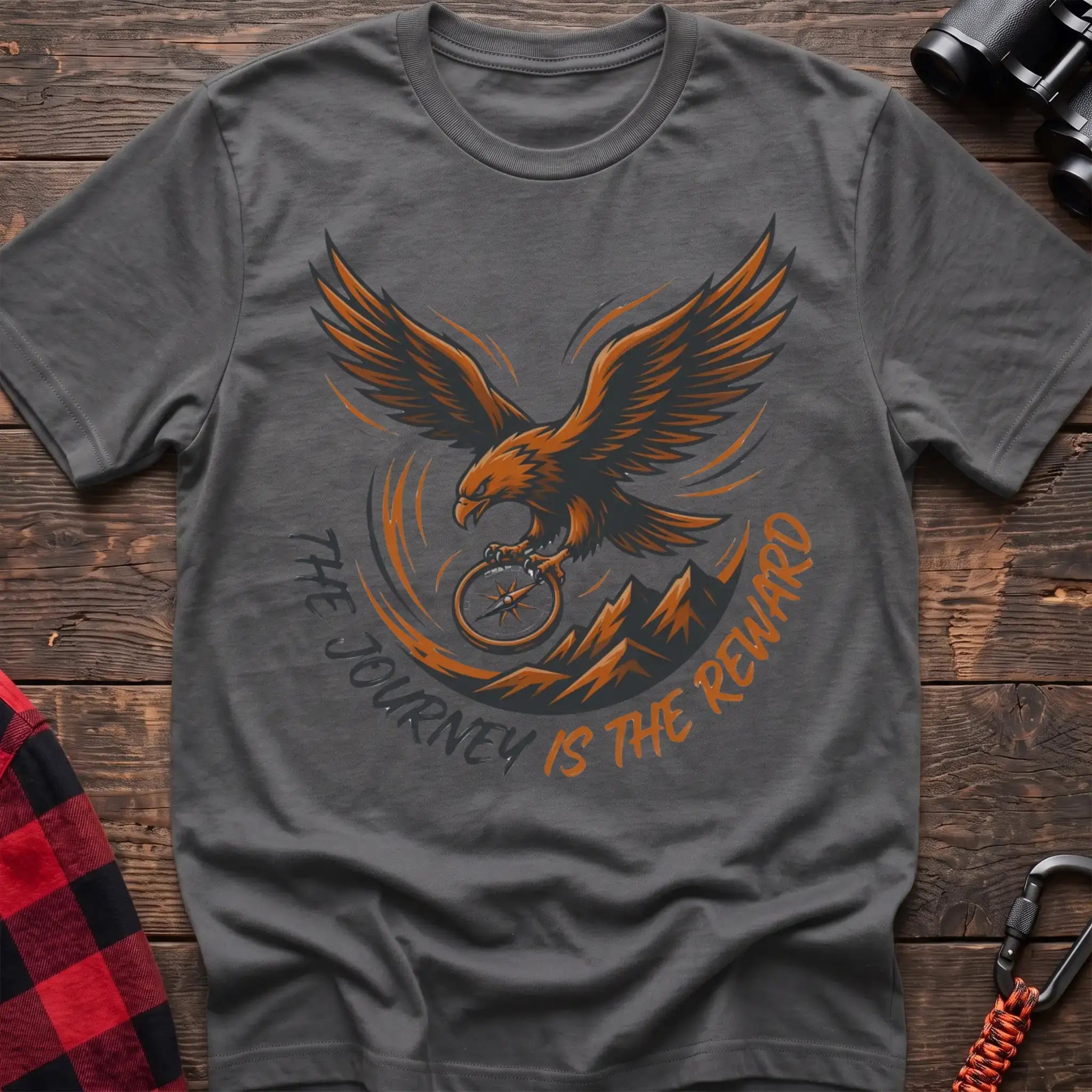 Graphite heather t-shirt with an orange and dark graphic of a soaring eagle holding a compass above mountains, with the curved text “THE JOURNEY IS THE REWARD,” laid flat on a wooden surface.