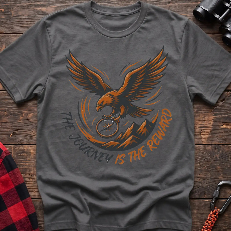 Graphite heather t-shirt with an orange and dark graphic of a soaring eagle holding a compass above mountains, with the curved text “THE JOURNEY IS THE REWARD,” laid flat on a wooden surface.