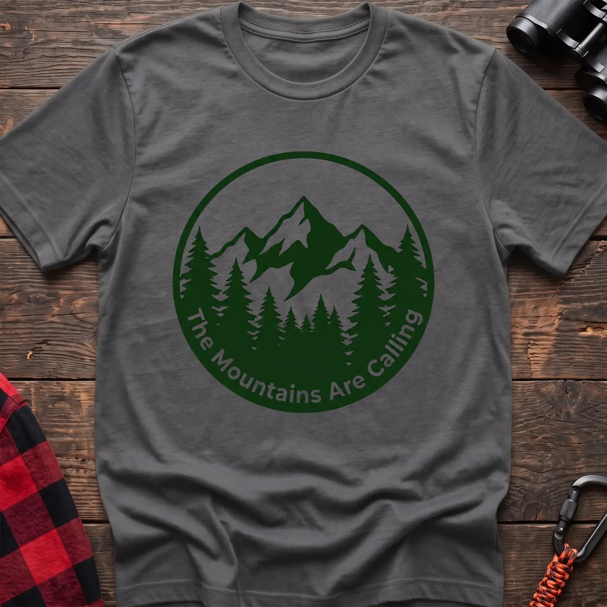 Graphite heather t-shirt with a circular dark green design featuring mountain peaks and pine trees, with the text “The Mountains Are Calling,” laid flat on a wooden surface.