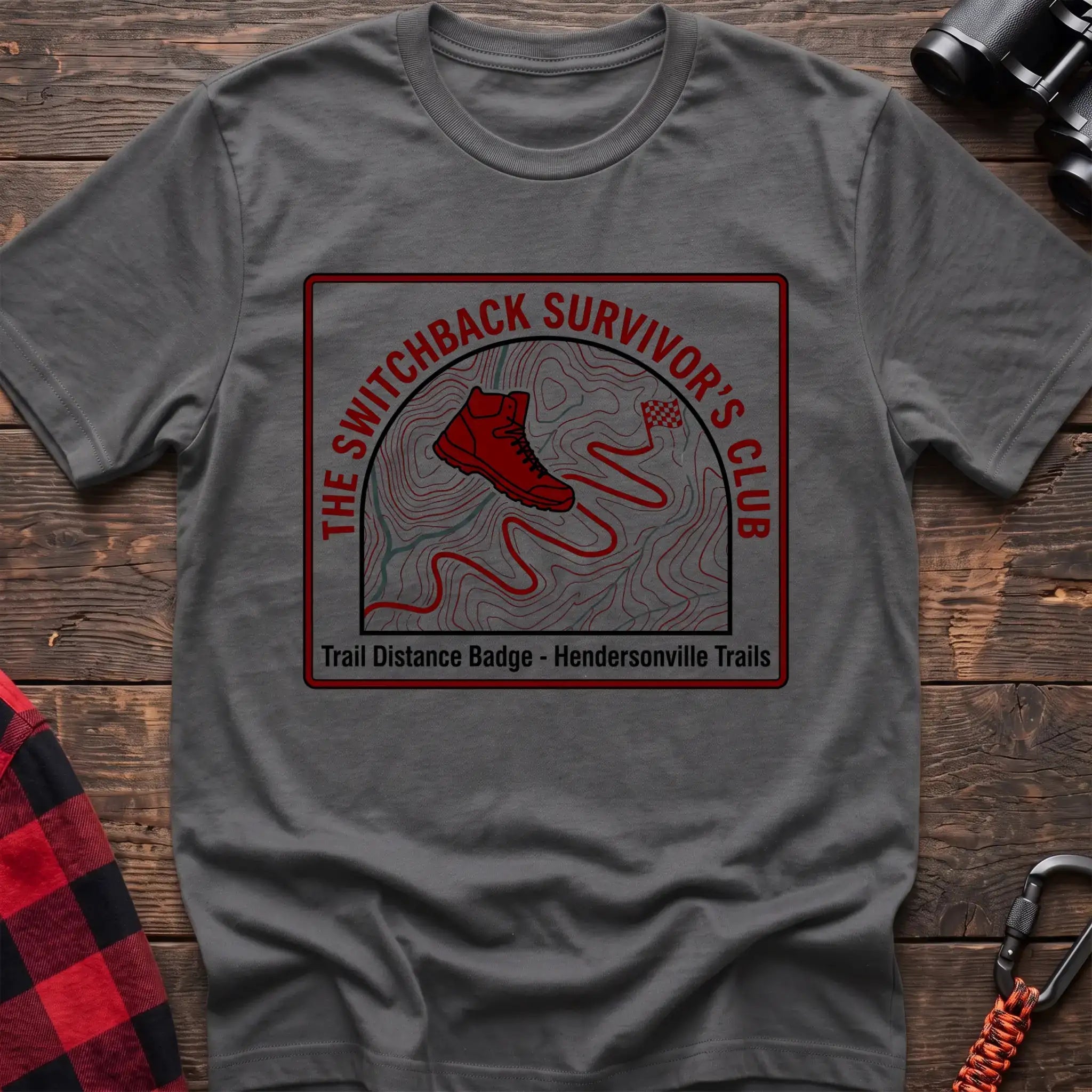 Graphite heather t-shirt with a square badge design featuring a red hiking boot on a topographic map with a winding trail, and the text “THE SWITCHBACK SURVIVOR’S CLUB” and “Trail Distance Badge - Hendersonville Trails,” laid flat on a wooden surface.