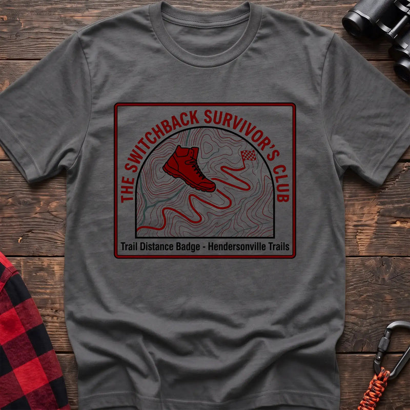 Graphite heather t-shirt with a square badge design featuring a red hiking boot on a topographic map with a winding trail, and the text “THE SWITCHBACK SURVIVOR’S CLUB” and “Trail Distance Badge - Hendersonville Trails,” laid flat on a wooden surface.