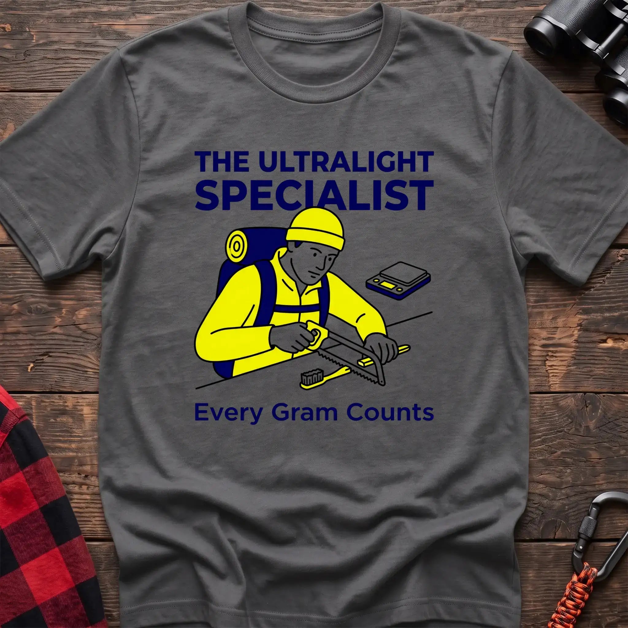 Graphite heather t-shirt with an illustration of a hiker sawing a toothbrush on a table next to a scale, with the text “THE ULTRALIGHT SPECIALIST” and “Every Gram Counts,” laid flat on a wooden surface.