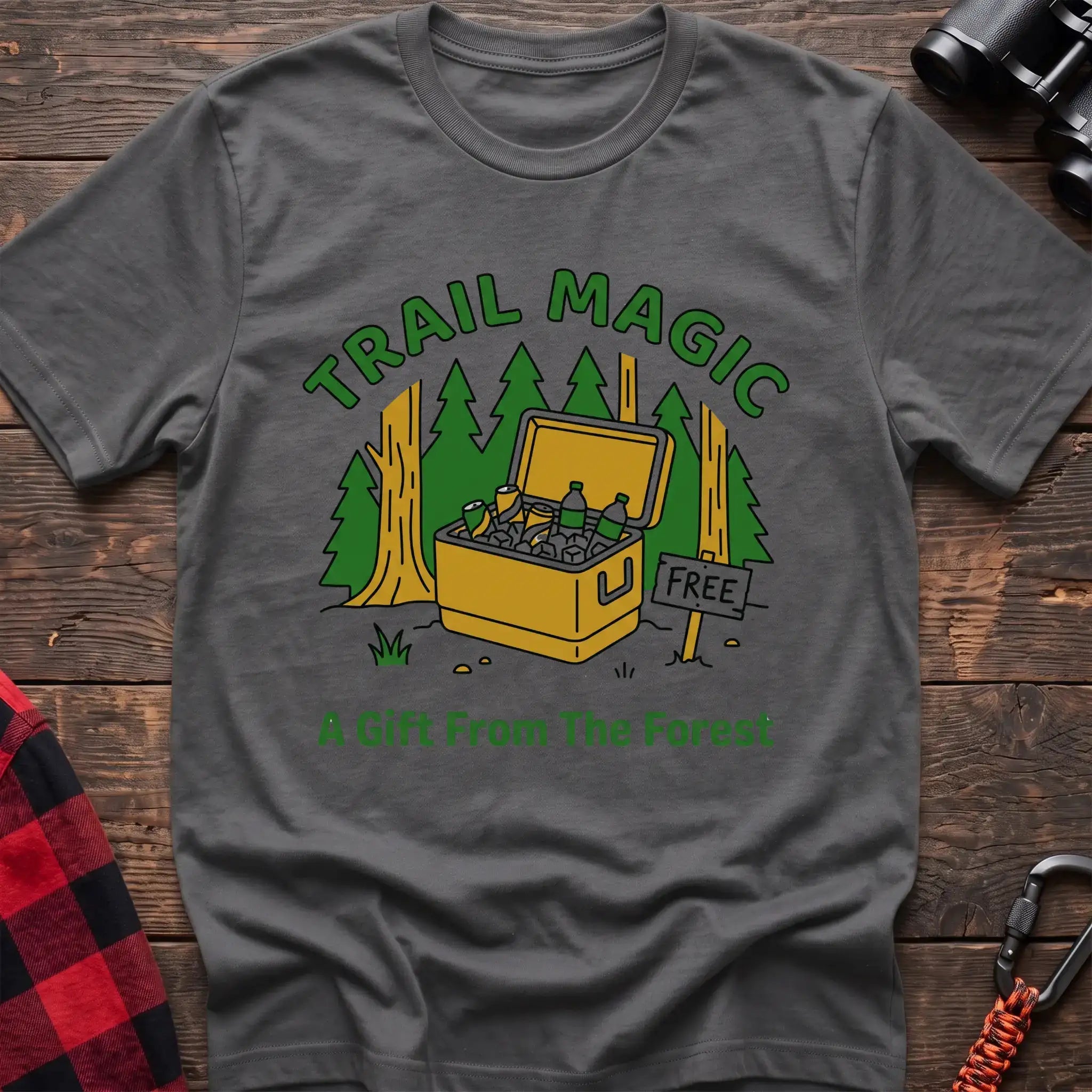 Graphite heather t-shirt with an illustration of an open cooler filled with drinks in a forest setting, with the text “TRAIL MAGIC” and “A Gift From The Forest,” laid flat on a wooden surface.