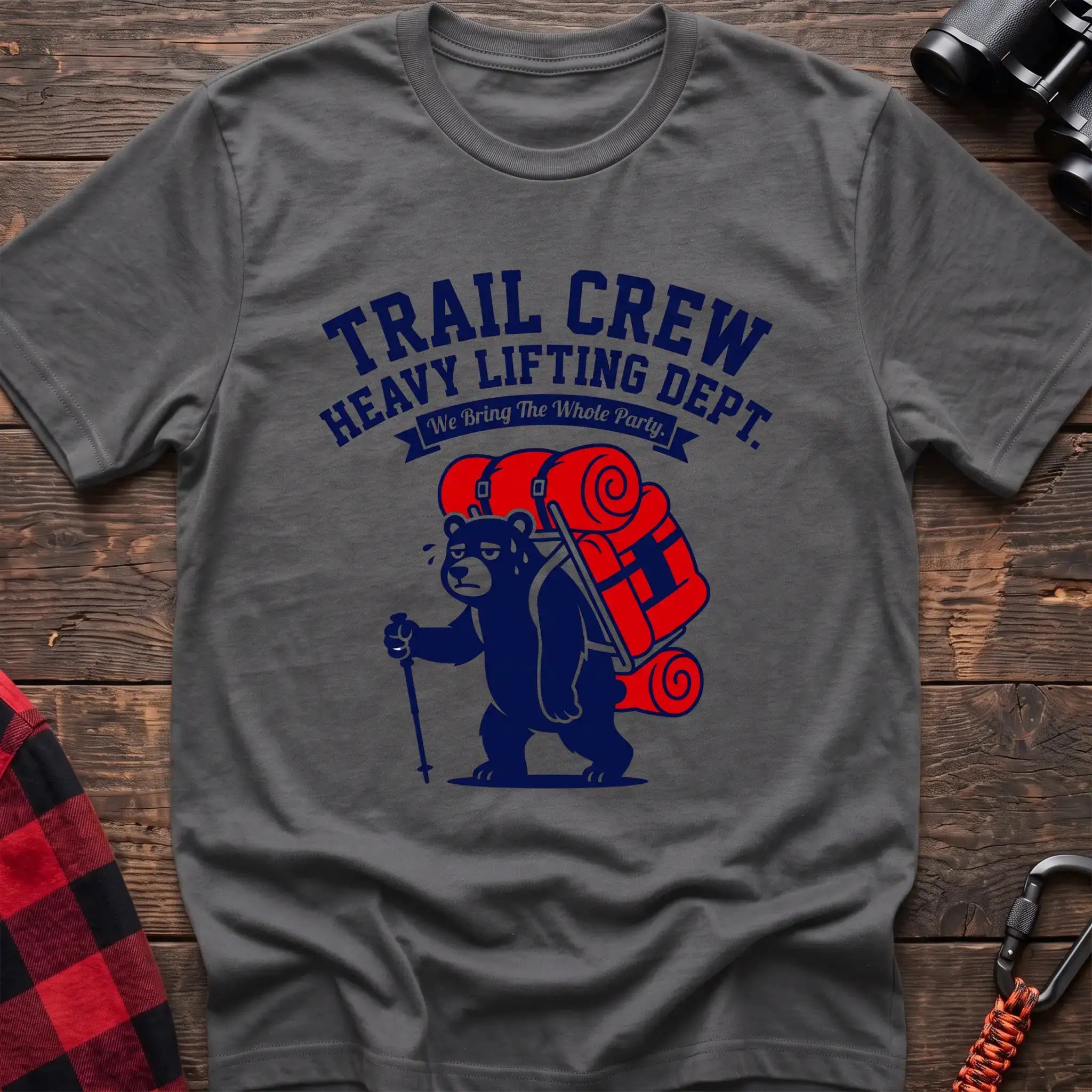 Graphite heather t-shirt with a cartoon dark blue bear carrying a large red backpack and hiking pole, with the text “TRAIL CREW HEAVY LIFTING DEPT. We Bring The Whole Party,” laid flat on a wooden surface.