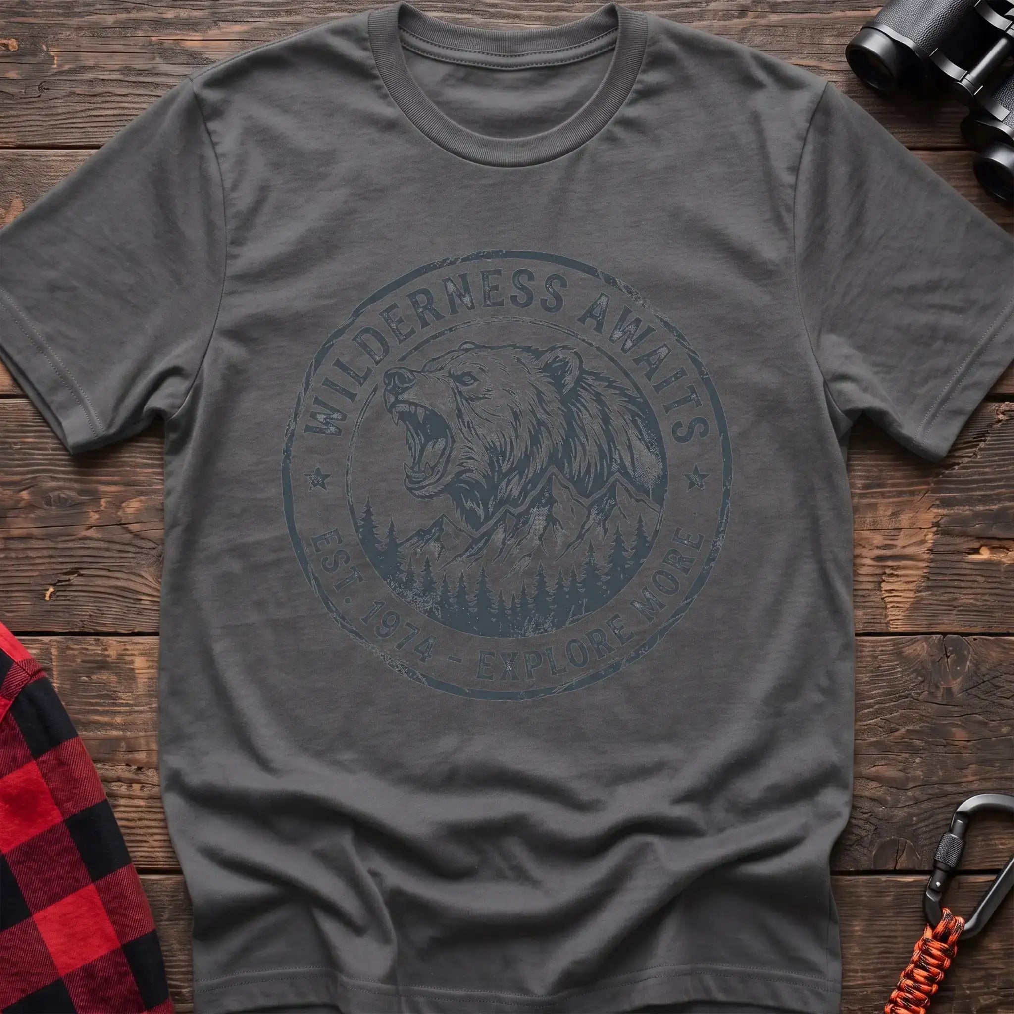 Graphite heather t-shirt with a circular dark gray badge design featuring a roaring bear head above mountains and pine trees, with the text “WILDERNESS AWAITS” and “EST. 1974 - EXPLORE MORE,” laid flat on a wooden surface.