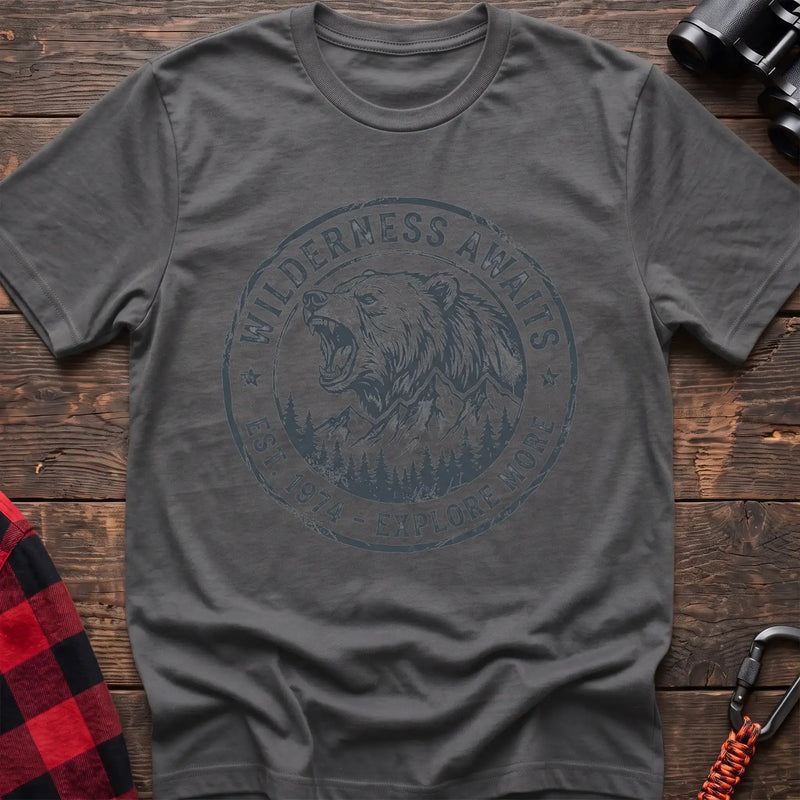 Graphite heather t-shirt with a circular dark gray badge design featuring a roaring bear head above mountains and pine trees, with the text “WILDERNESS AWAITS” and “EST. 1974 - EXPLORE MORE,” laid flat on a wooden surface.