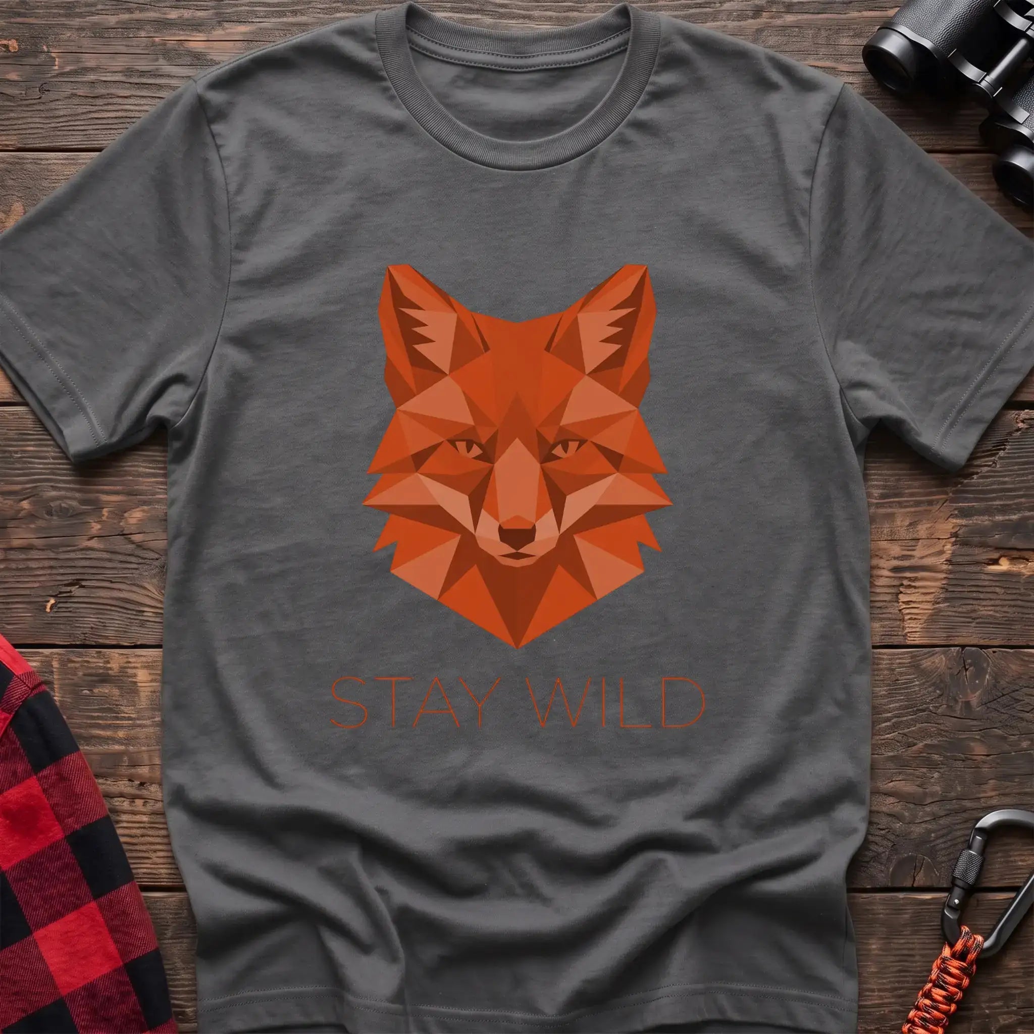 Graphite heather t-shirt with a geometric low-poly orange fox head and the text “STAY WILD” printed below in matching orange, laid flat on a wooden surface.