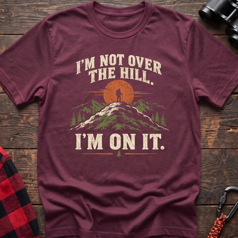 Heather maroon cotton t-shirt with a hiker on a summit graphic and I’m Not Over the Hill I’m On It text on rustic wood