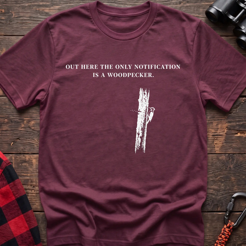 Heather maroon cotton t-shirt with a woodpecker graphic and Out Here the Only Notification Is a Woodpecker text on rustic wood
