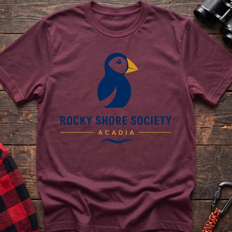 Heather maroon cotton t-shirt with a navy puffin logo, yellow beak, and Rocky Shore Society Acadia text on a rustic wood background
